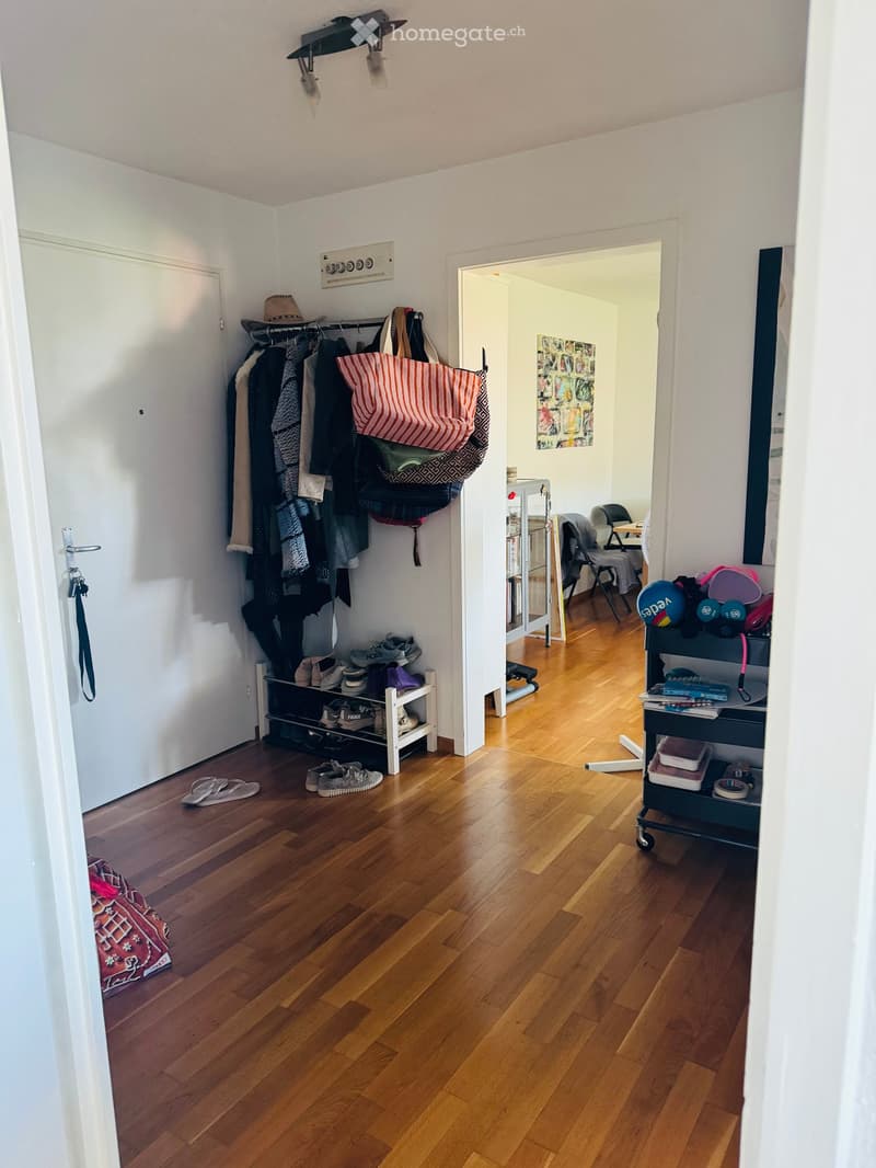 A narrow hallway with wooden flooring, a coat rack with jackets and bags, a shoe shelf, and a small cart with items. The hallway leads to another room with a chair and a table.
