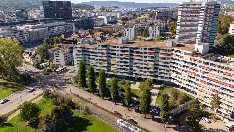 Aerial view of a modern apartment complex with colorful facades, surrounded by trees, roads, and other buildings in an urban setting.