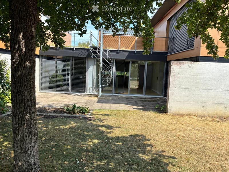 Modern house exterior with a spiral staircase leading to a rooftop terrace, large glass windows, and a small garden area with grass and a tree.