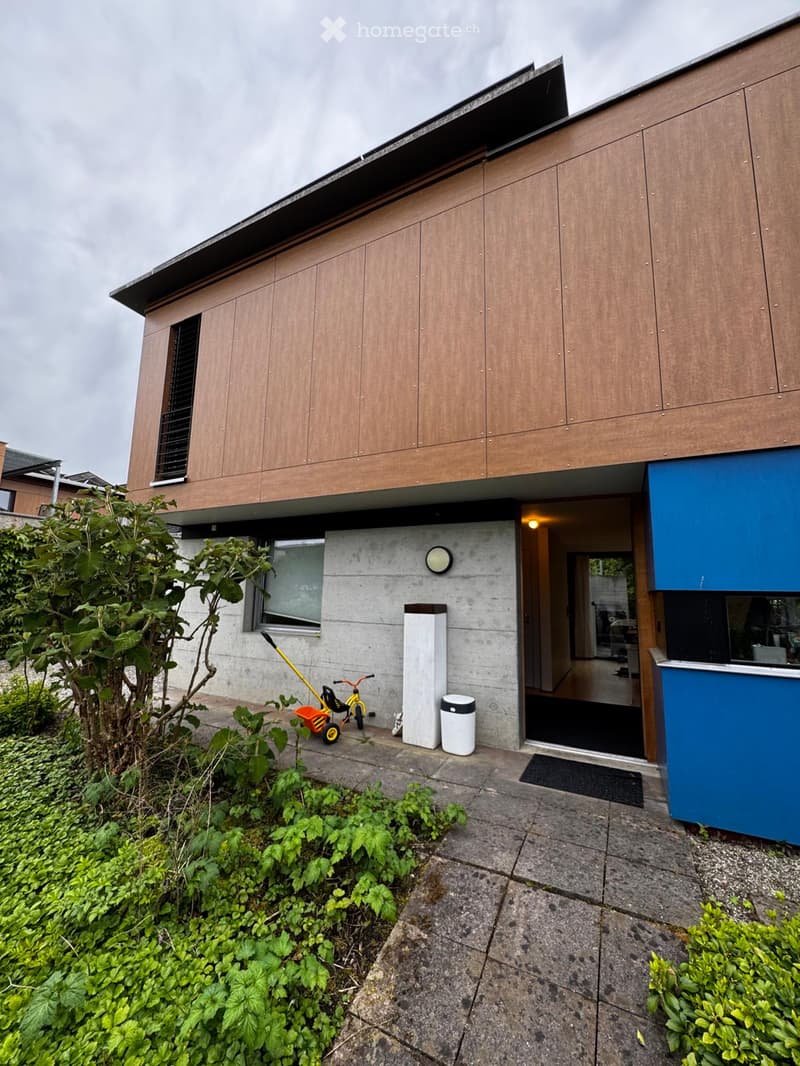 Modern two-story house with wooden and concrete exterior, featuring a small garden area with plants and a bicycle, and a visible entrance with a blue accent wall.