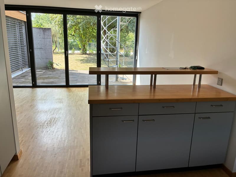 Modern kitchen with grey cabinets, wooden countertop, and a small table with keys and a wallet on it. Large sliding glass doors lead to an outdoor area.