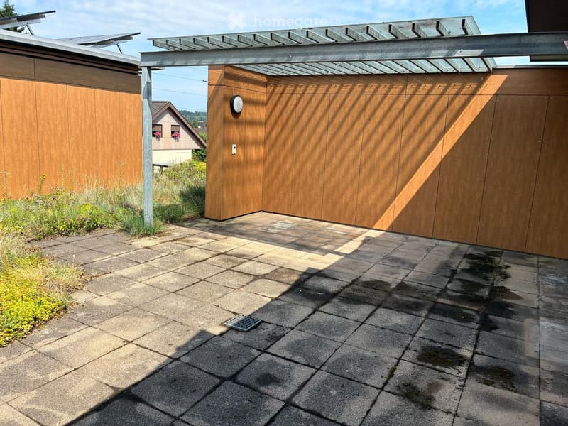 Outdoor patio area with stone tiles, wooden shed, covered pergola, and small garden plants.