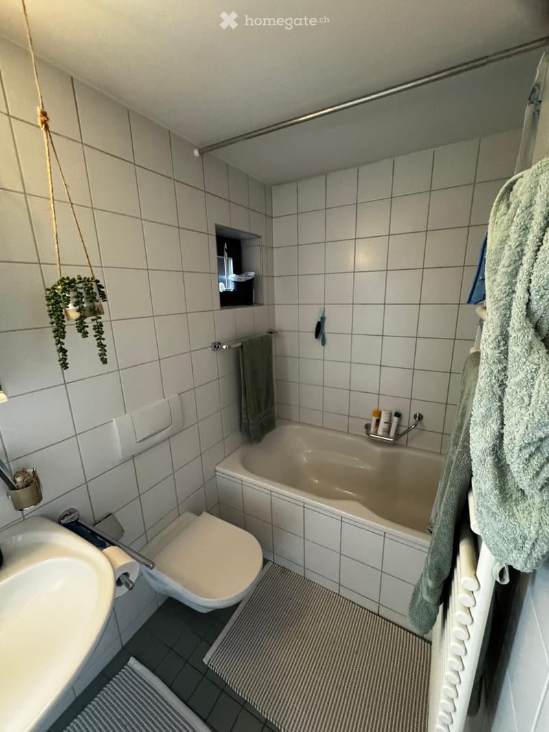 A small bathroom with white tiled walls and floor, featuring a bathtub, toilet, sink, and towels hanging on the side.