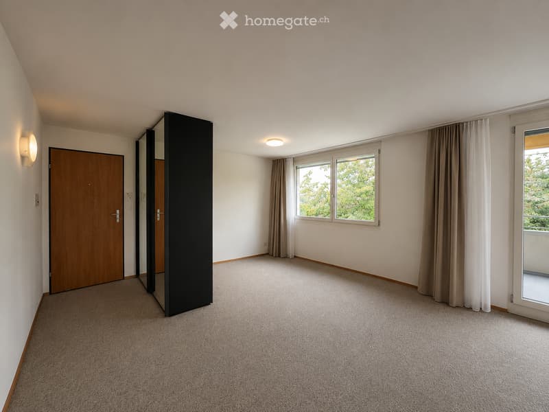 Empty room with light-colored walls, carpeted floor, large windows with curtains, and a sliding glass door leading to a balcony.