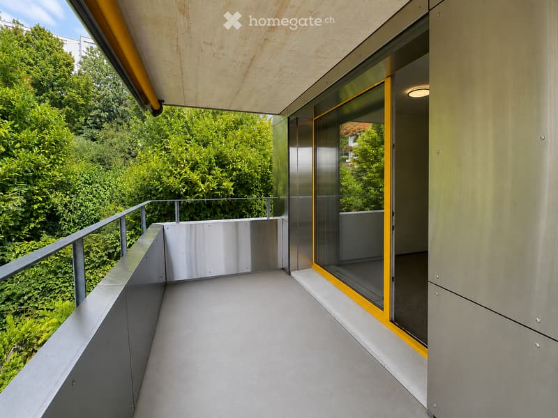 Modern balcony with a sleek metal railing, glass walls, and a wooden ceiling, overlooking a lush green garden.