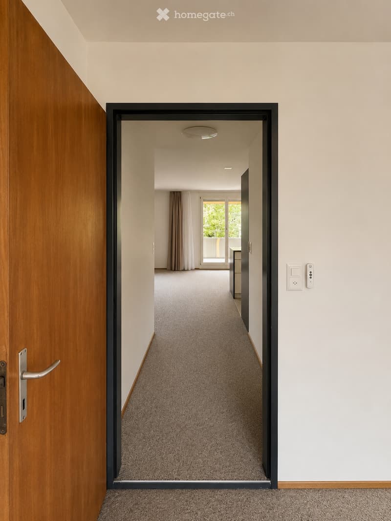A modern, carpeted hallway with light-colored walls, a wooden door on the left, and an open doorway leading to another room with a view of a balcony and greenery outside.