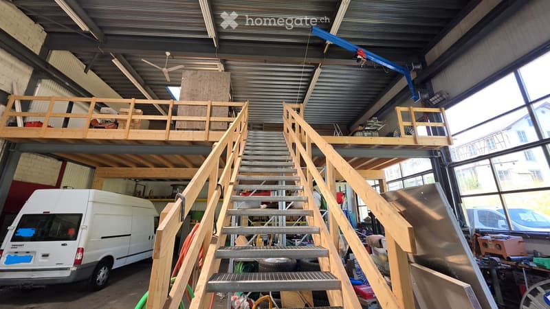 Large industrial-style garage with a metal roof, wooden staircase leading to an upper loft area, and a white van parked inside.