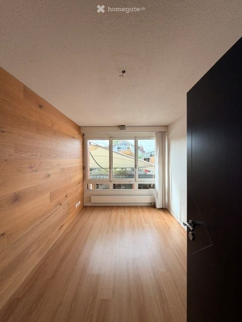 Empty room with wooden flooring, white walls, and large windows with white frames. The room has a black door on one side and a radiator below the windows.
