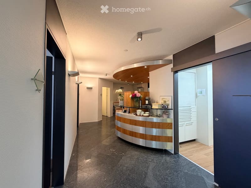 Modern reception area with a curved wooden and metallic reception desk, vases with flowers, coffee machine, and framed picture on the desk.