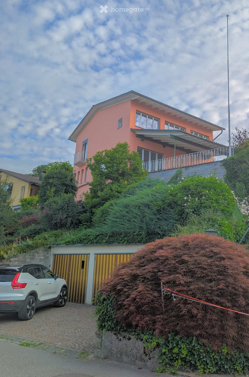 Two-story house with pink exterior, white-framed windows, and a balcony on the upper level. Garage with yellow striped doors is visible, along with a parked car.