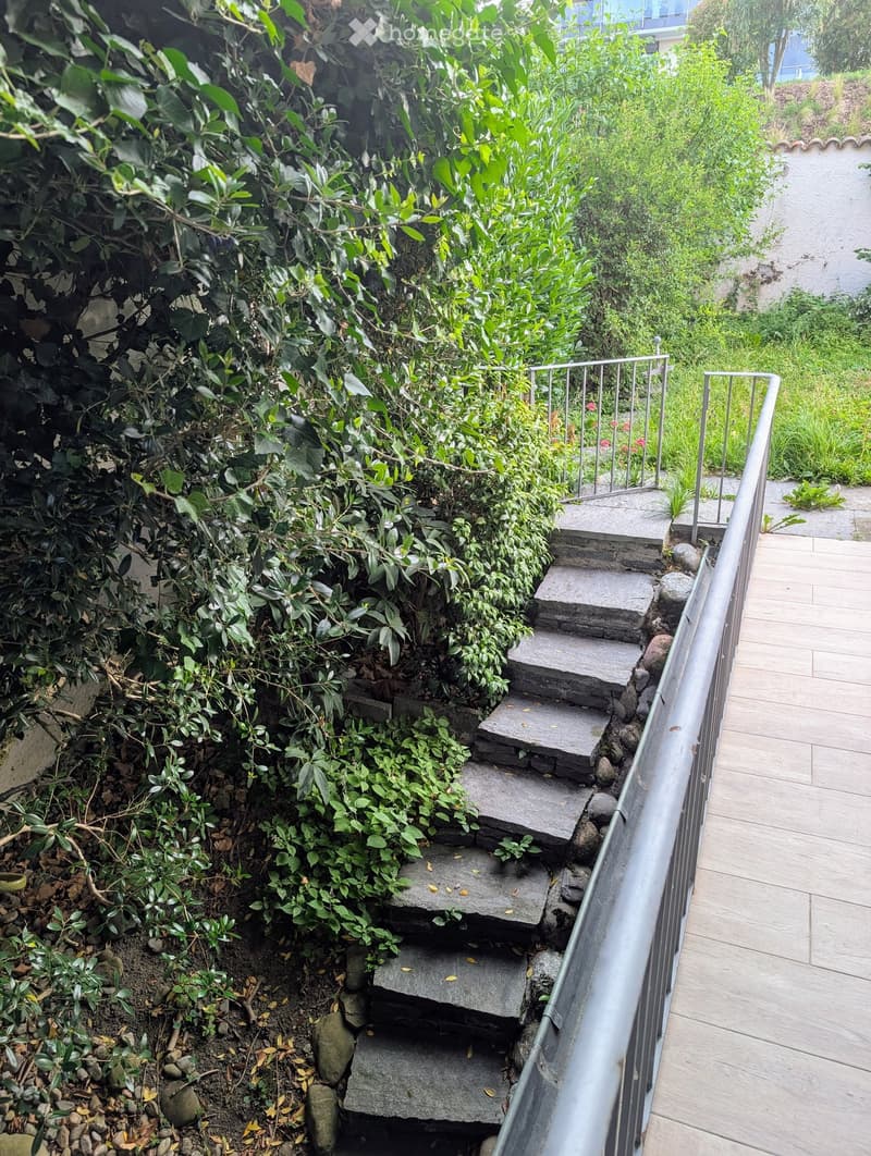 A narrow, elevated garden area with stone steps leading down to a lower level, surrounded by lush greenery and shrubs.