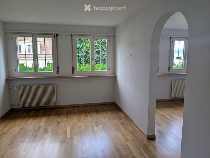 Empty room with wooden flooring, white walls, large windows, radiators, and an arched doorway leading to another space.