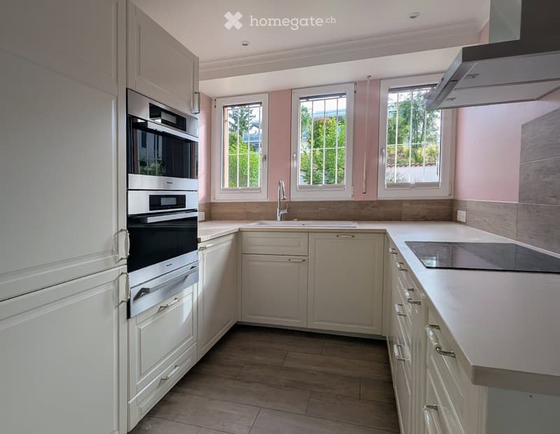 Modern kitchen with white cabinets, stainless steel oven, central island with countertop, and three windows offering natural light.