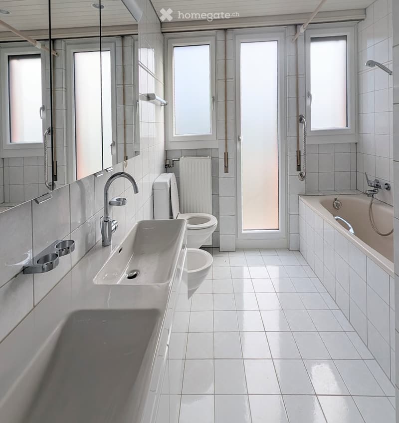 Modern bathroom with white tiles, a large sink, toilet, bathtub, and a radiator. The space is bright with multiple windows and a large mirror.