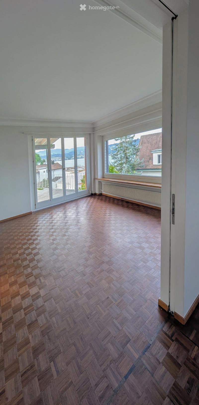 Empty room with wooden herringbone flooring, white walls, large windows, and sliding glass doors leading to a balcony.