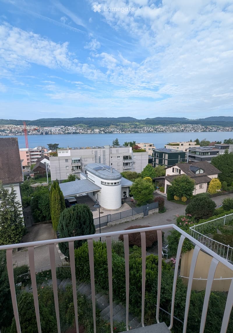 Balcony overlooking a residential area with modern buildings, greenery, and a scenic view of a lake and distant mountains.
