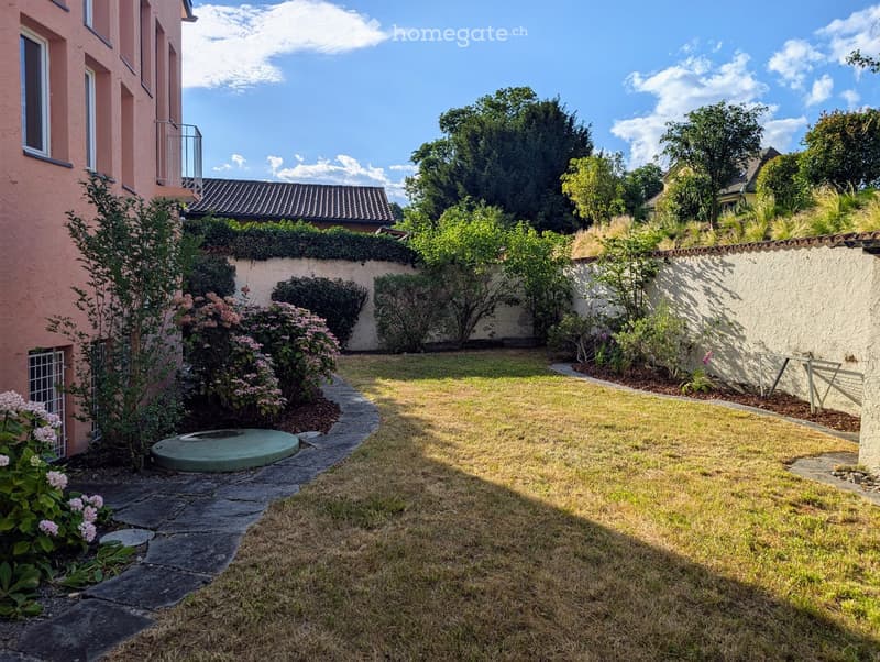 Well-maintained garden with a stone pathway, circular stone platform, various shrubs and flowering plants, and a low hedge along the wall.