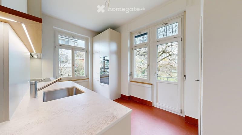 Modern kitchen with white cabinets, a stainless steel sink, and a marble countertop, featuring large windows and a red-tiled floor.