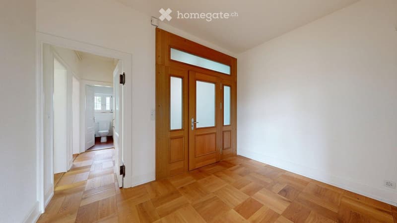 Empty hallway with wooden flooring, white walls, and a large wooden double door with glass panels leading to another room.