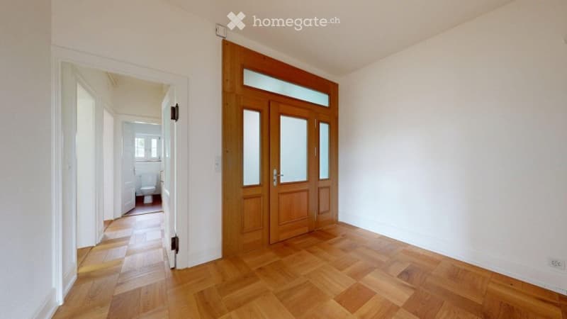 Empty room with wooden flooring and white walls, featuring a large wooden door with glass panels leading to another room.