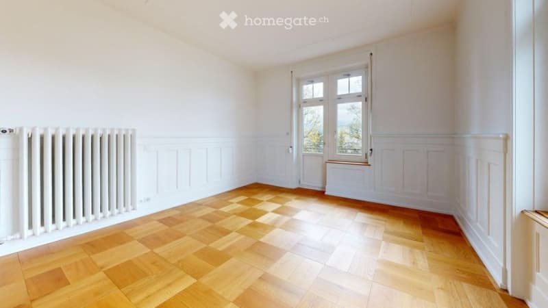 Empty room with wooden flooring, white walls, and a radiator. Large window with a view of greenery outside.