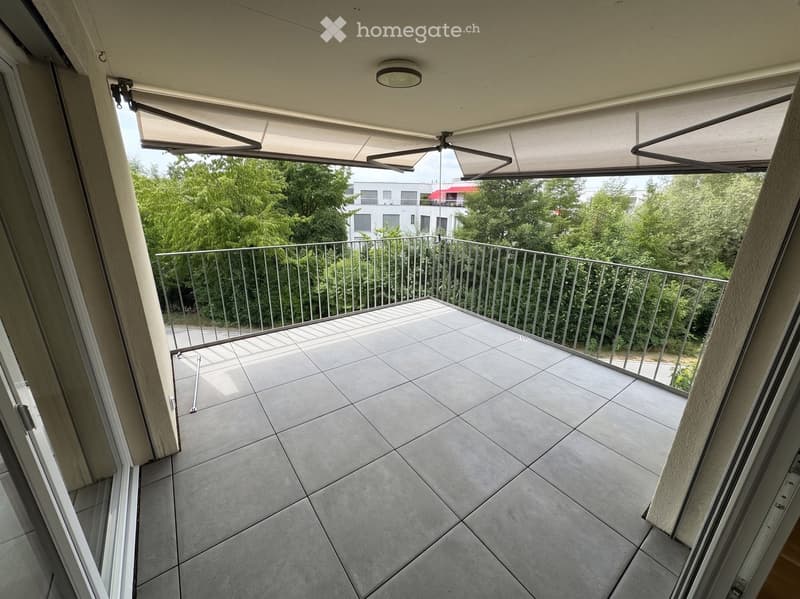 Balcony with tiled floor, metal railing, retractable awning, ceiling light, and view of greenery and buildings.