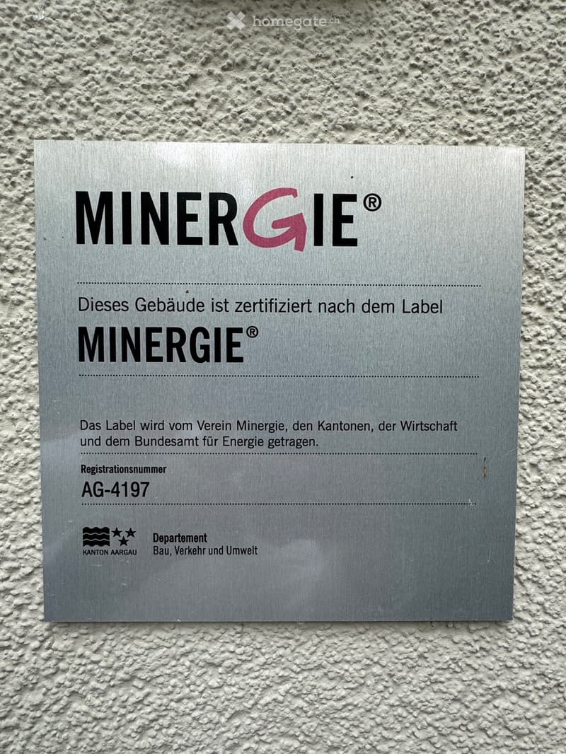 A silver plaque mounted on a textured wall, displaying the Minergie certification logo and details about the building's energy certification.
