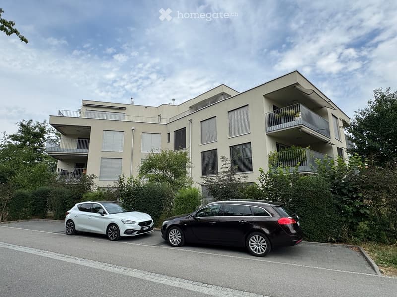 Modern multi-story residential building with balconies, surrounded by greenery and parked cars.