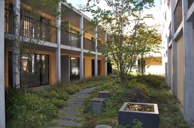 Courtyard area with a pathway, small seating areas, and modern buildings with balconies and glass windows.