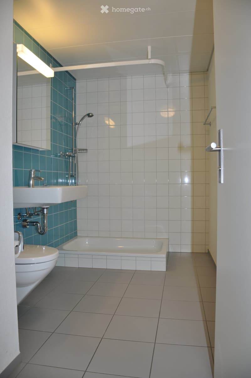 Modern bathroom with white tiles, a bathtub, toilet, sink, and a mirror above the sink. The walls feature a combination of white and blue tiles.