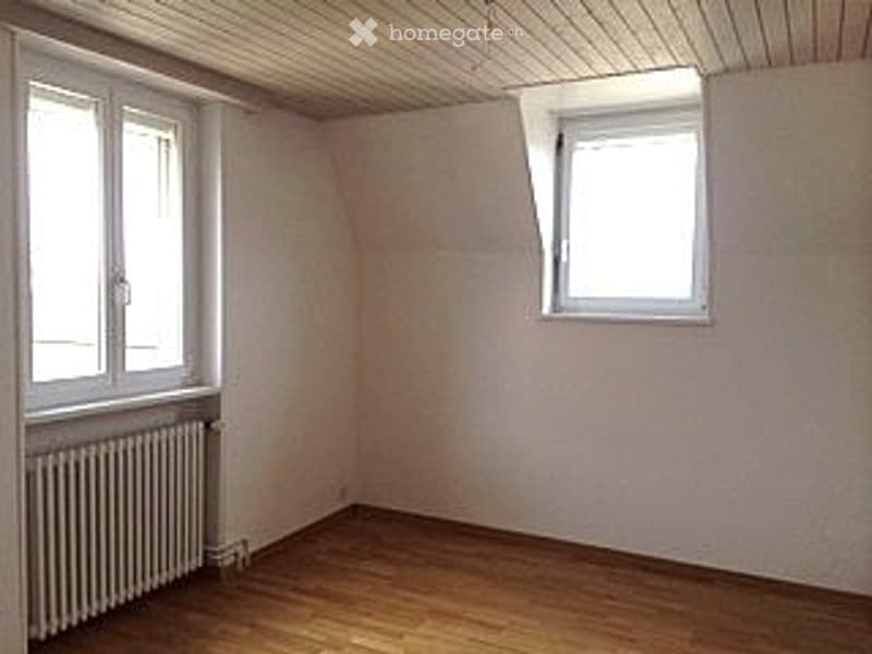 Empty room with wooden flooring, white walls, and two windows, one with a slanted wall.