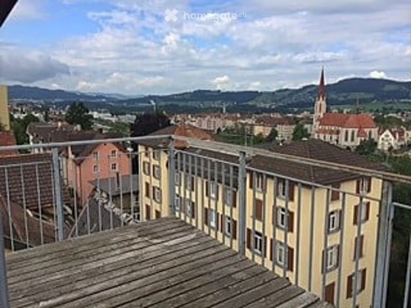 Balcony with wooden decking, metal railing, overlooking a town with buildings and distant mountains.