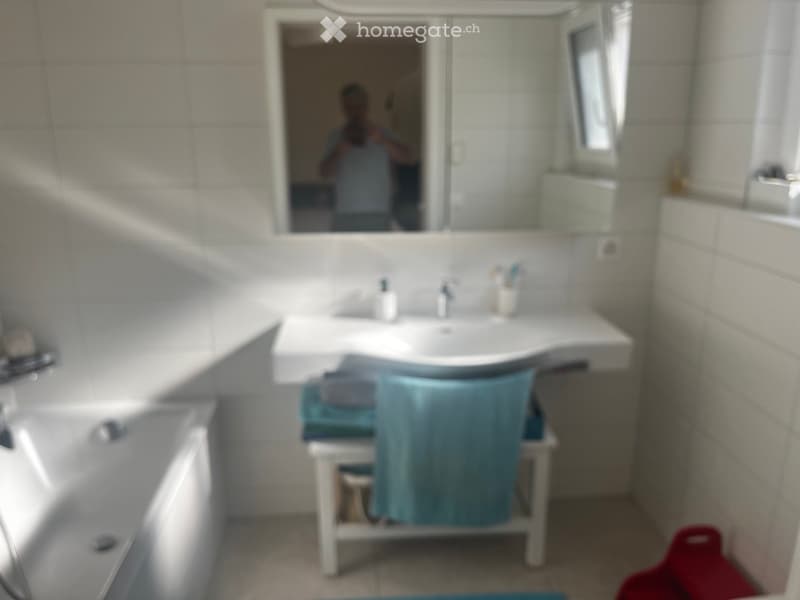 Modern bathroom with a white sink, mirror, and bathtub. Blue towels are draped over a small white stand next to the sink.