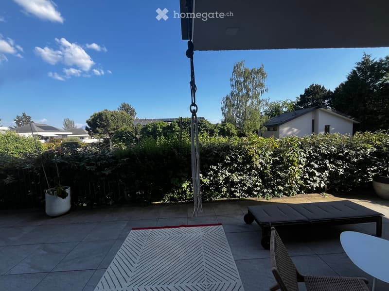 Outdoor patio with tiled floor, white table, wicker chair, dark bench, and a hanging decoration.