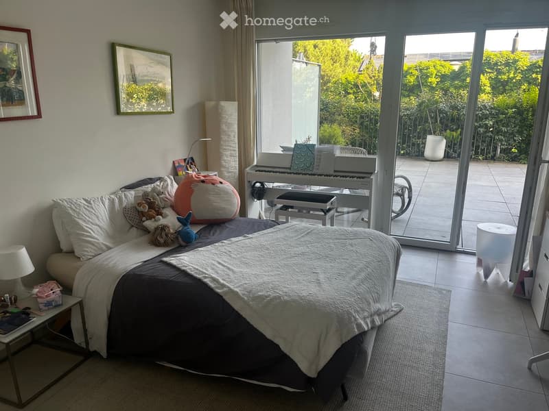Cozy bedroom with a large bed, sofa, small table, lamp, and piano near a large window with a view of greenery.