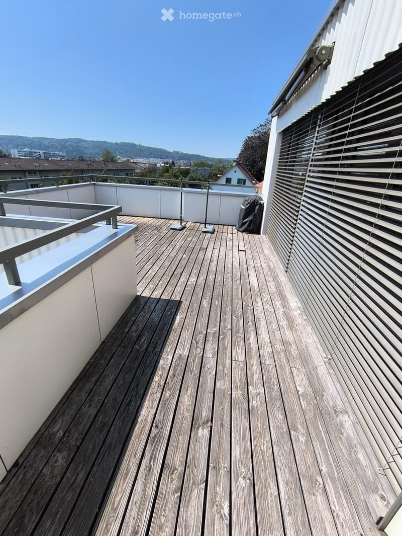 Wooden deck with metal railing, white walls, and a view of the city and hills in the background.