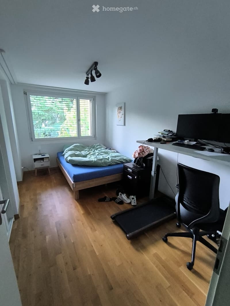 A furnished room with a bed, desk, chair, screen, keyboard, laptop, nightstand, picture frame, treadmill, drawer, and lamp.