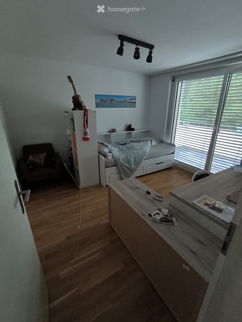 A modern bedroom with a bed, a chair, a guitar on a shelf, a table with various items, and a large window with blinds.