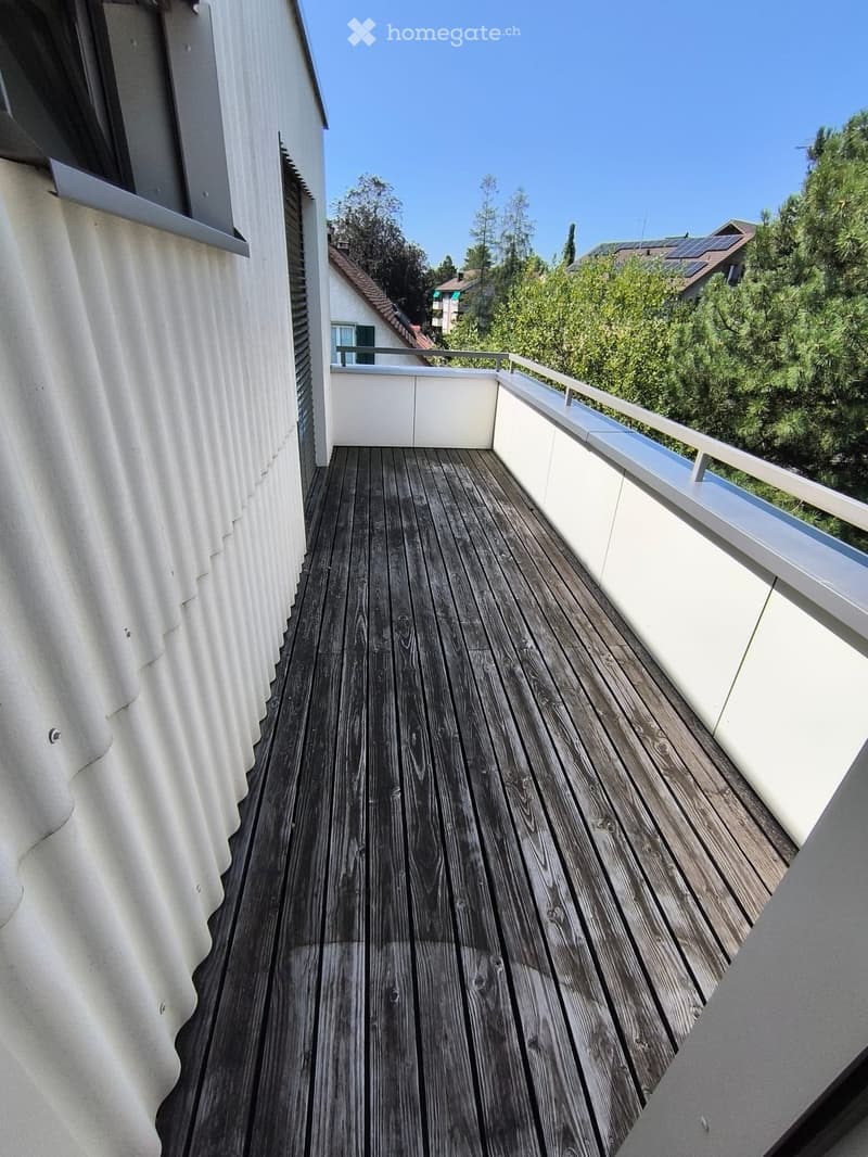 Wooden deck with white railings, overlooking a residential area with trees and solar panels on neighboring roofs.
