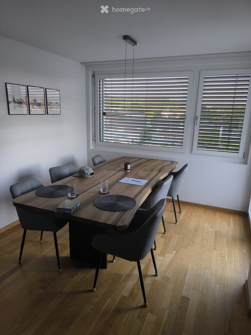 Modern dining area with a long wooden table, six chairs, and various items on the table including a cup, placemats, a book, and a hat. Large windows with blinds and a pendant light fixture are visible.