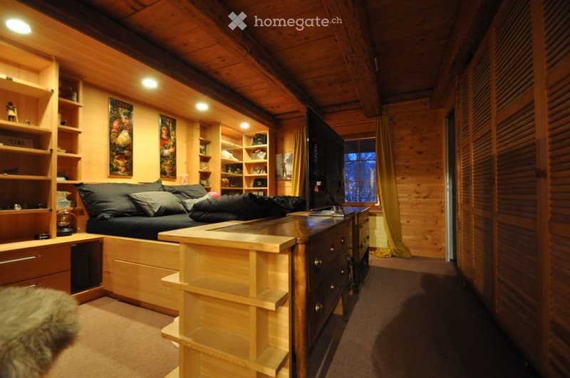 Cozy wooden bedroom with a large sofa, wooden table, television, shelves, and a lamp. The room features exposed wooden beams and warm lighting.