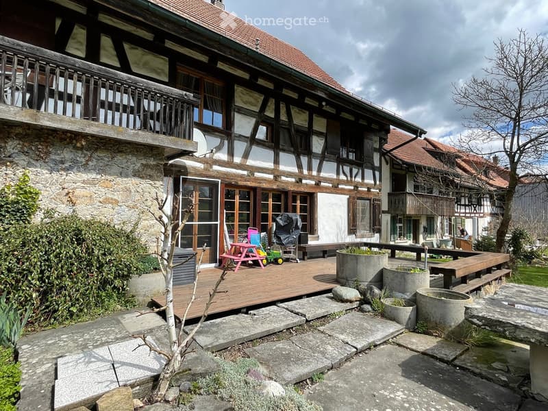 Wooden deck area attached to a traditional house, featuring a picnic table, a small toy car, a bench, and several large concrete pots with plants.