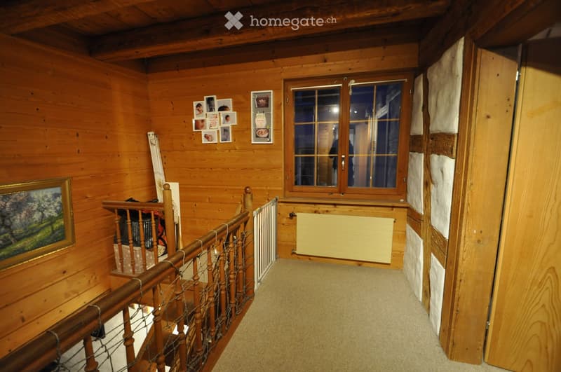 Wooden hallway with a staircase, wooden walls, a window, and a radiator. The staircase features wooden railings and a metal safety gate at the bottom.