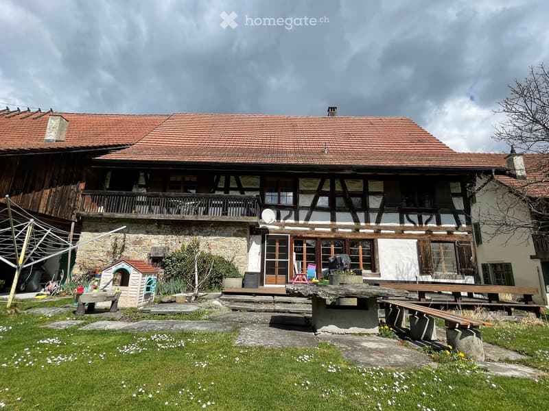 Two-story house with red-tiled roof, stone and wooden facade, large windows, and a balcony. Outdoor seating area with benches and a picnic table.