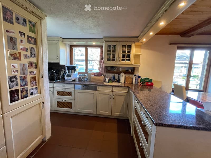 Bright kitchen with white cabinets, granite countertop, and stainless steel appliances including a refrigerator, dishwasher, and coffee maker. The space features a large window and a dining area with a table and chairs.