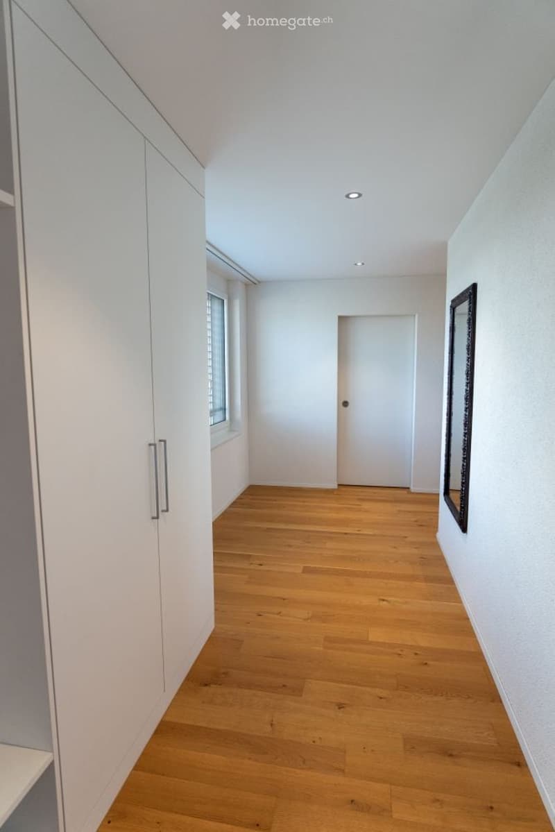 A clean, modern hallway with light wooden flooring, white walls, built-in white cabinetry on one side, and a closed door at the end. A mirror is mounted on the wall to the right, and a small window allows natural light to enter.