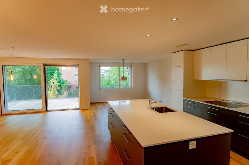 Modern kitchen with light-colored countertops, dark wood cabinets, and a central island. Features include a sink, faucet, and built-in stove. Large windows and sliding glass doors provide natural light and access to the outdoor area.