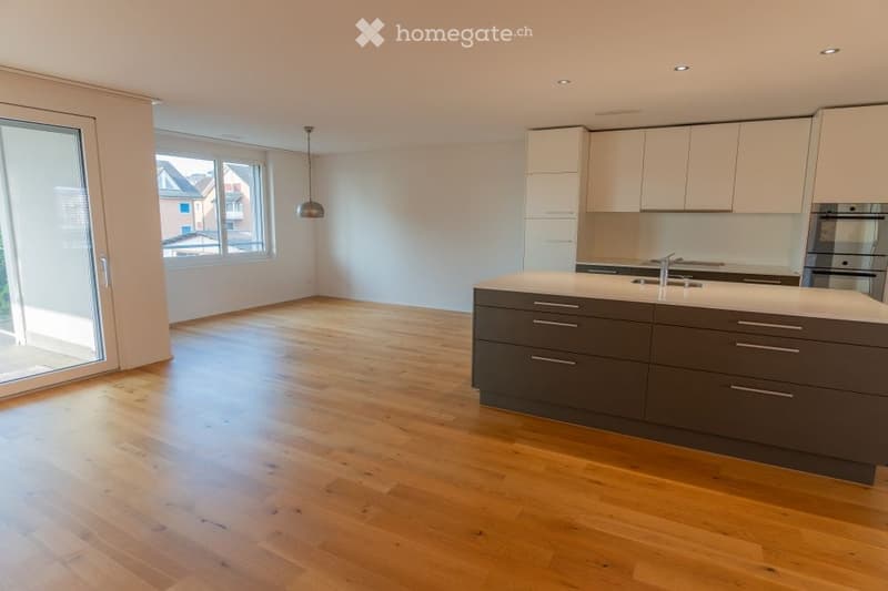 Empty kitchen with wooden flooring, white cabinets, and a central island. Large windows and a sliding glass door provide natural light.