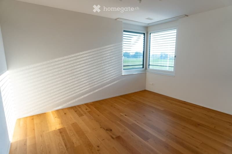 Empty room with light wooden flooring and white walls, featuring large windows with blinds and natural light streaming in.