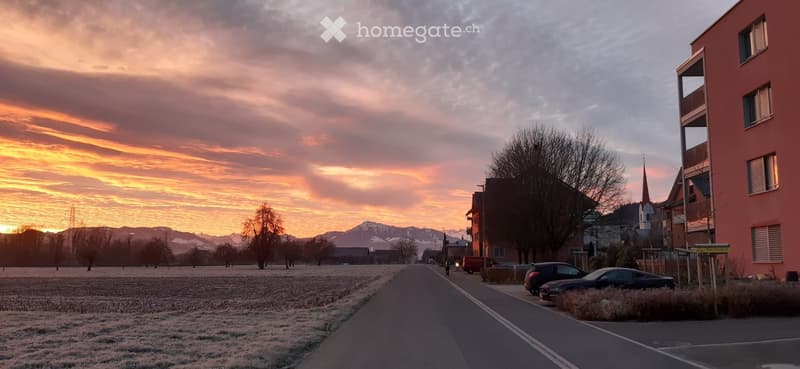 A serene town scene at sunset with snow-covered ground, a road leading towards buildings, and distant mountains.